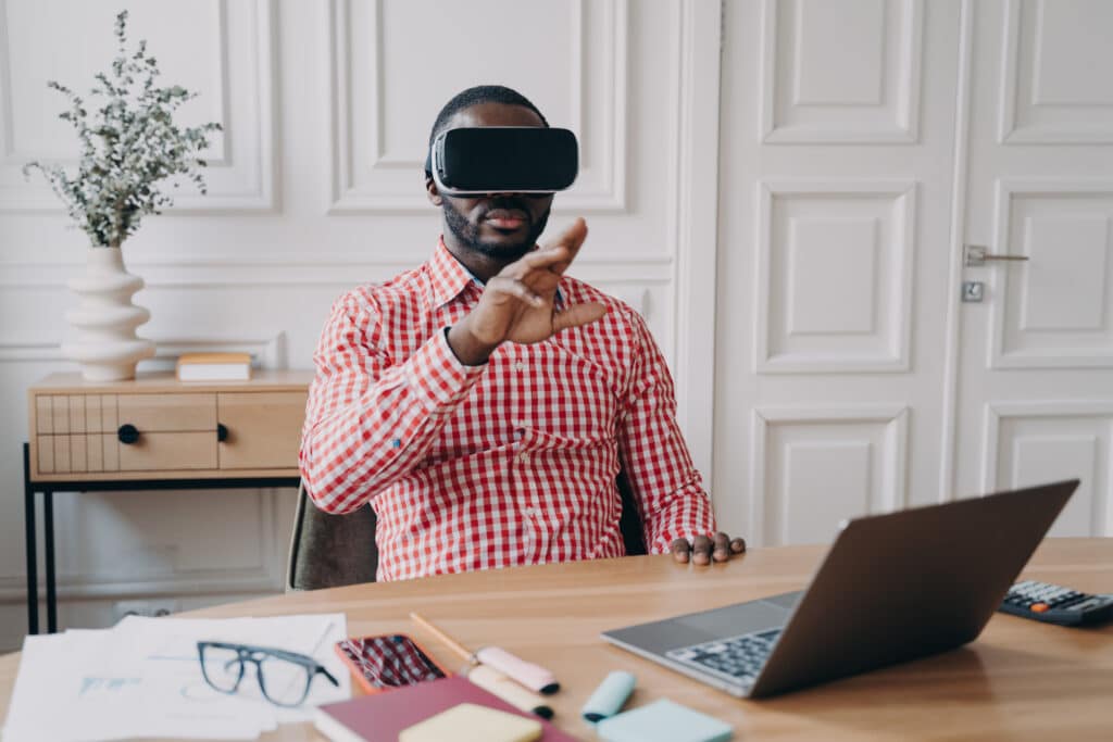 Young African ethnicity man entrepreneur in VR headset glasses immersed in 3D gaming in virtual reality relaxing during remote working online sitting at desk at modern home office in front of laptop